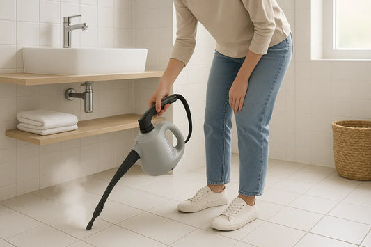 Adult woman using a handheld steam cleaner on grout between floor tiles in a bright modern bathroom with white and pale gray ceramic tiles